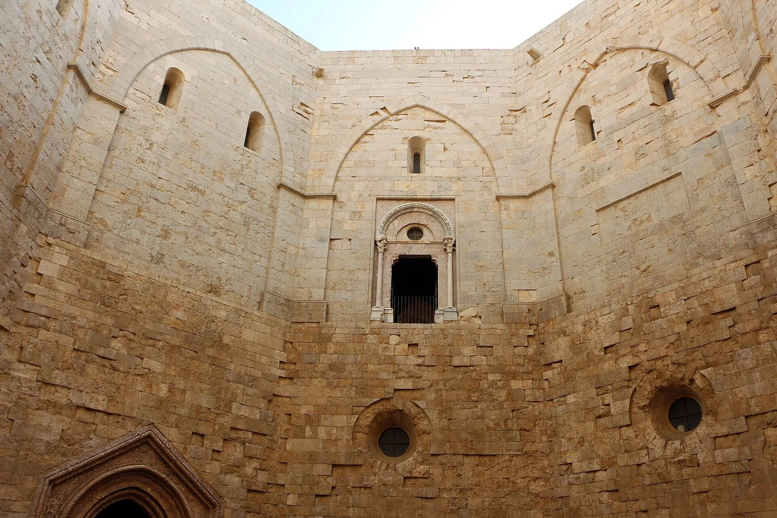 Il cortile interno di Castel del Monte visto dal basso: alte pareti in conci di pietra calcarea dorata, archi ciechi, un portale-finestra in marmo al piano superiore e oculi rotondi