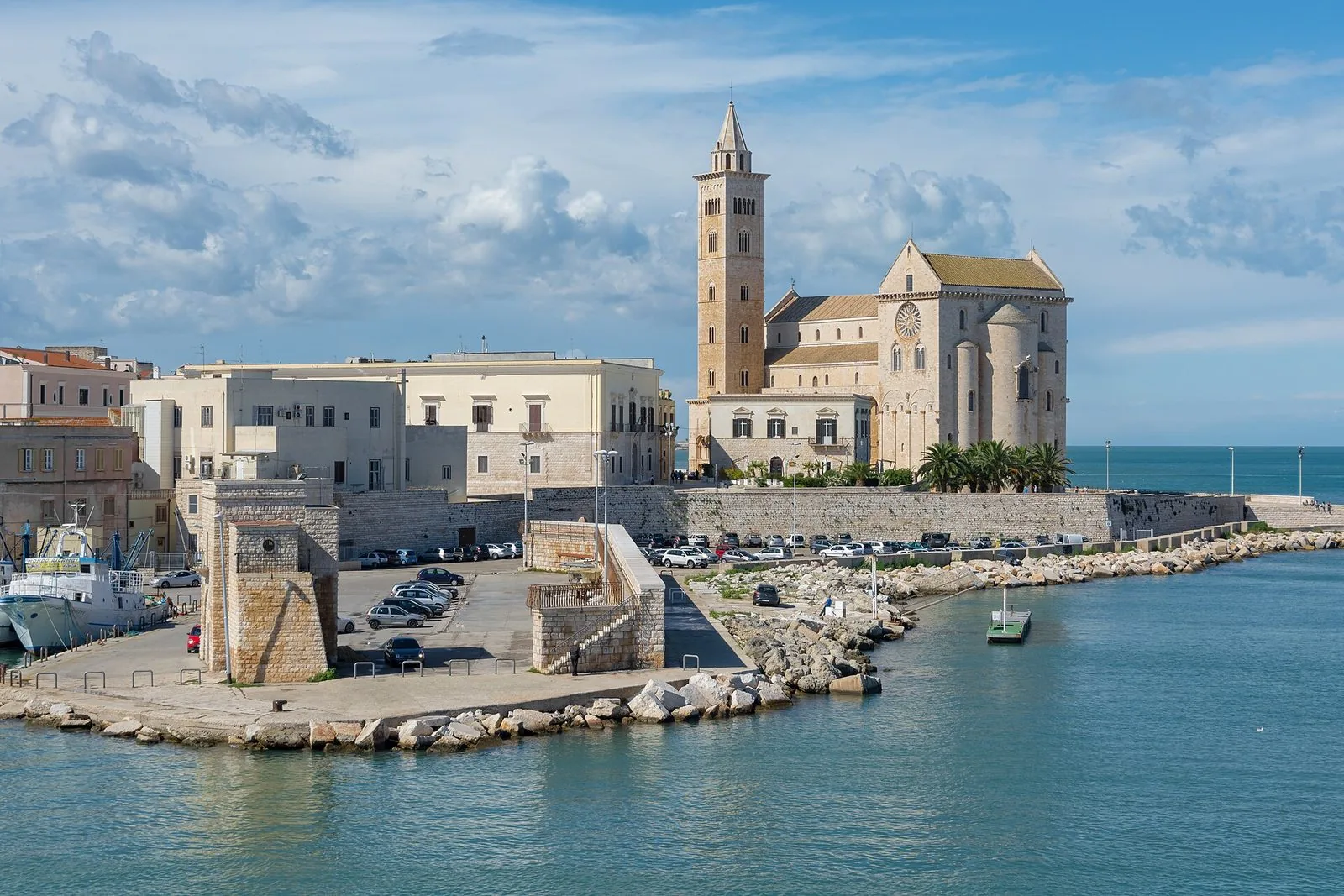 La Cattedrale di Trani con il campanile vista dal porto, con la banchina, un peschereccio ormeggiato a sinistra e l'acqua azzurra in primo piano
