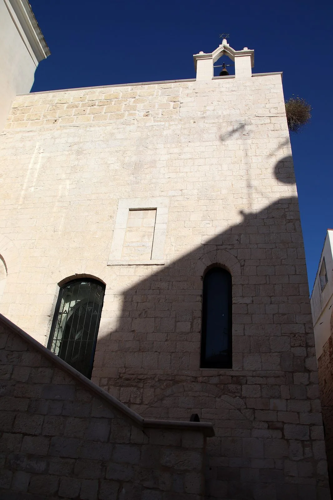 La Sinagoga Scolanova di Trani vista dal basso: alta parete in conci di pietra chiara, una scala esterna, finestre ad arco e un campanile a vela con la stella di David contro il cielo blu