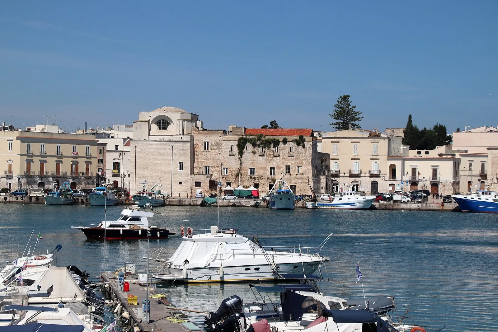 Il porto di Trani in una giornata limpida: barche ormeggiate in primo piano, pescherecci lungo la banchina e sullo sfondo la chiesa di Santa Teresa con la cupola, tra palazzi in pietra
