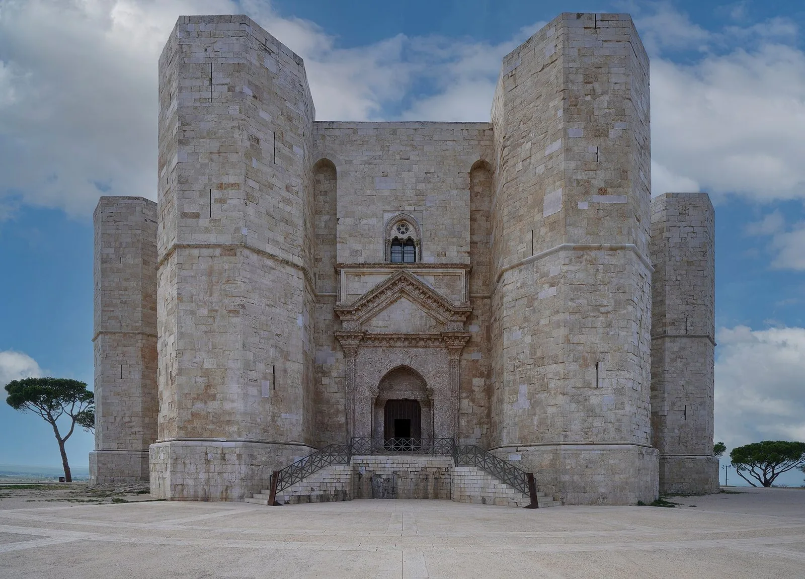 La facciata di Castel del Monte in pietra chiara con il portale d'ingresso, la scalinata e le torri ottagonali sotto un cielo azzurro con nuvole bianche